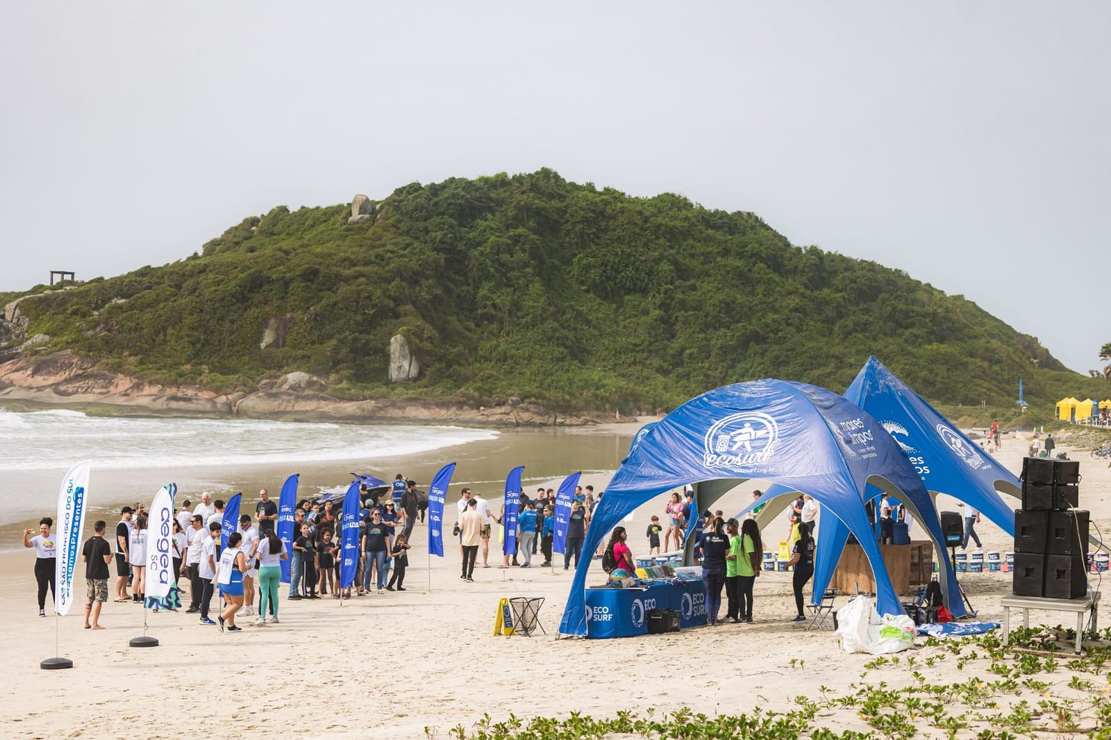 Voluntários reunidos na praia ao lado das tendas e bandeiras do Ecosurf, com o morro ao fundo