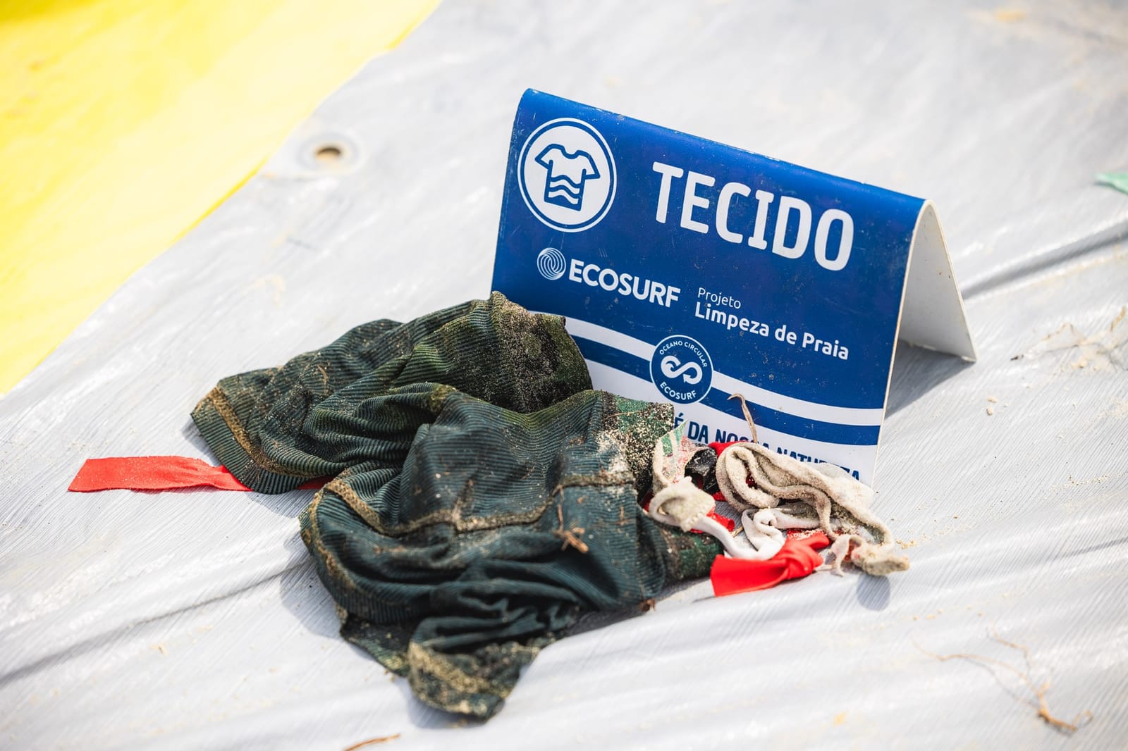 Tecidos recolhidos na praia ao lado da placa Tecido do projeto Limpeza de Praia