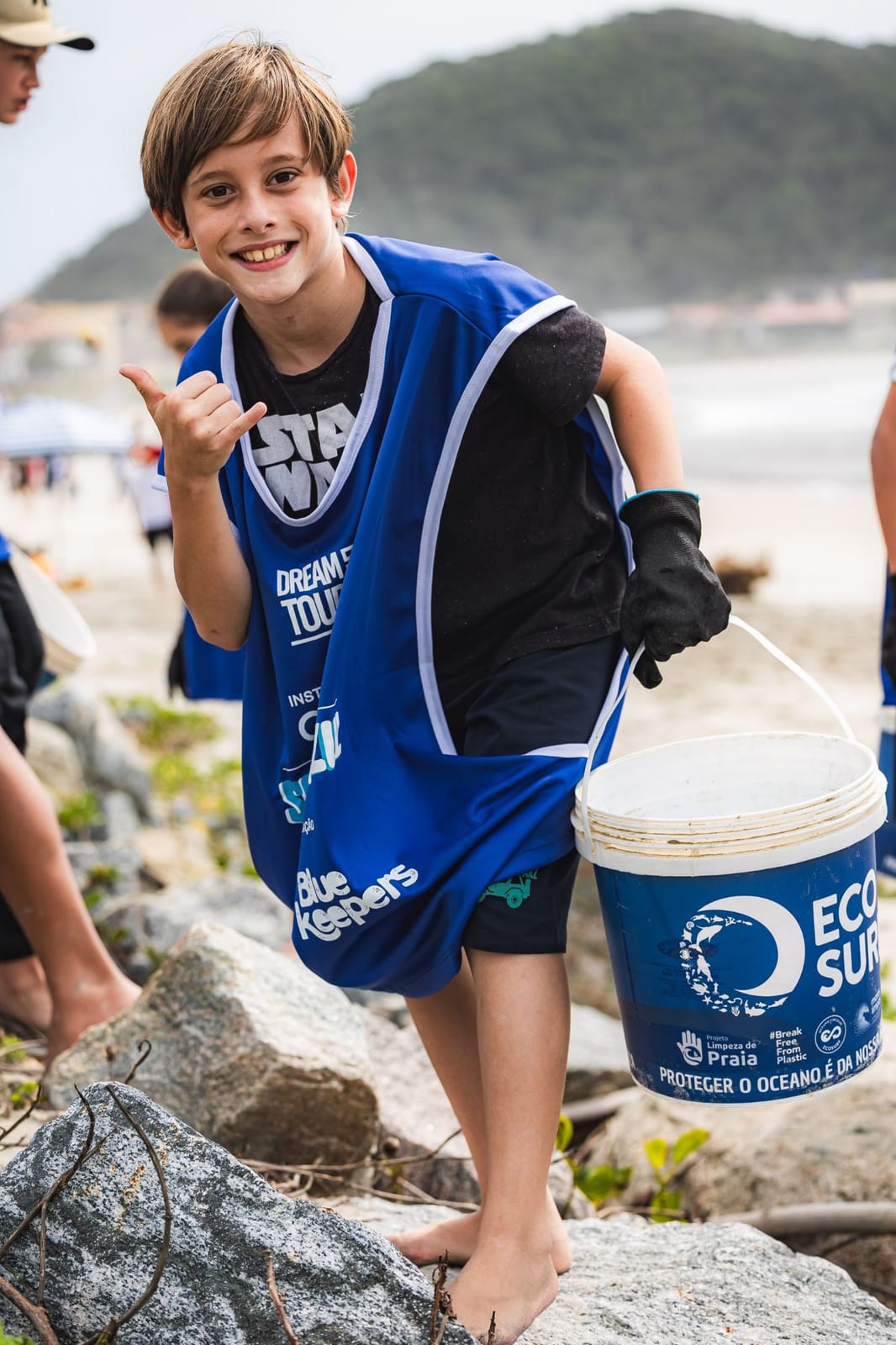 Menino voluntário sorri e faz o sinal de hang loose segurando um balde do Ecosurf