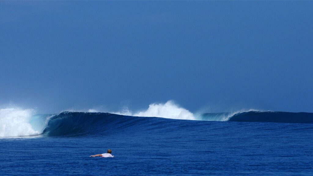 Surfista rema em direção a uma onda tubular em mar azul