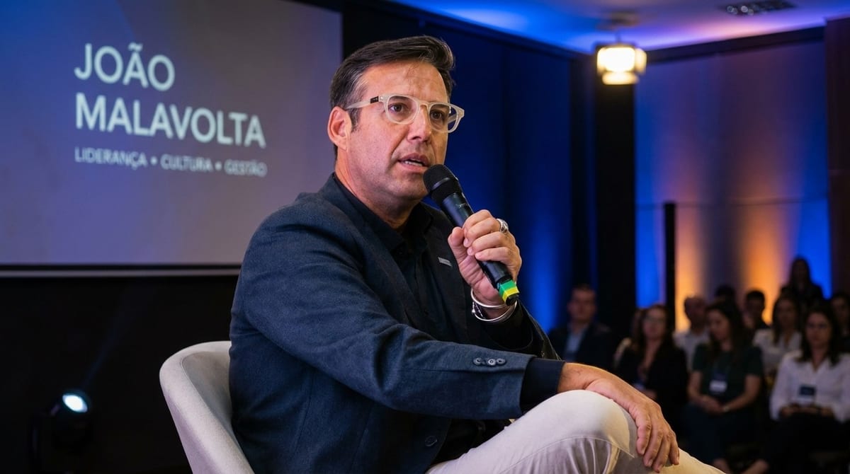 João Malavolta fala ao microfone sentado no palco, com seu nome projetado no telão