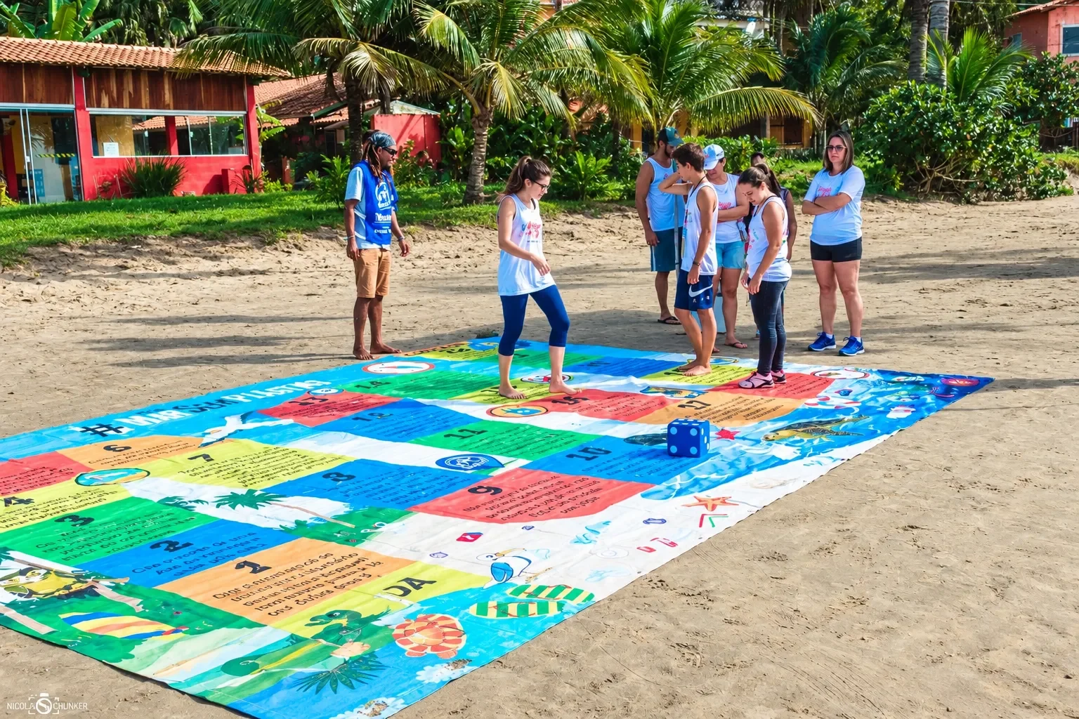 Grupo joga sobre o tabuleiro gigante Mar Sem Plástico na areia, com um dado azul sobre a lona