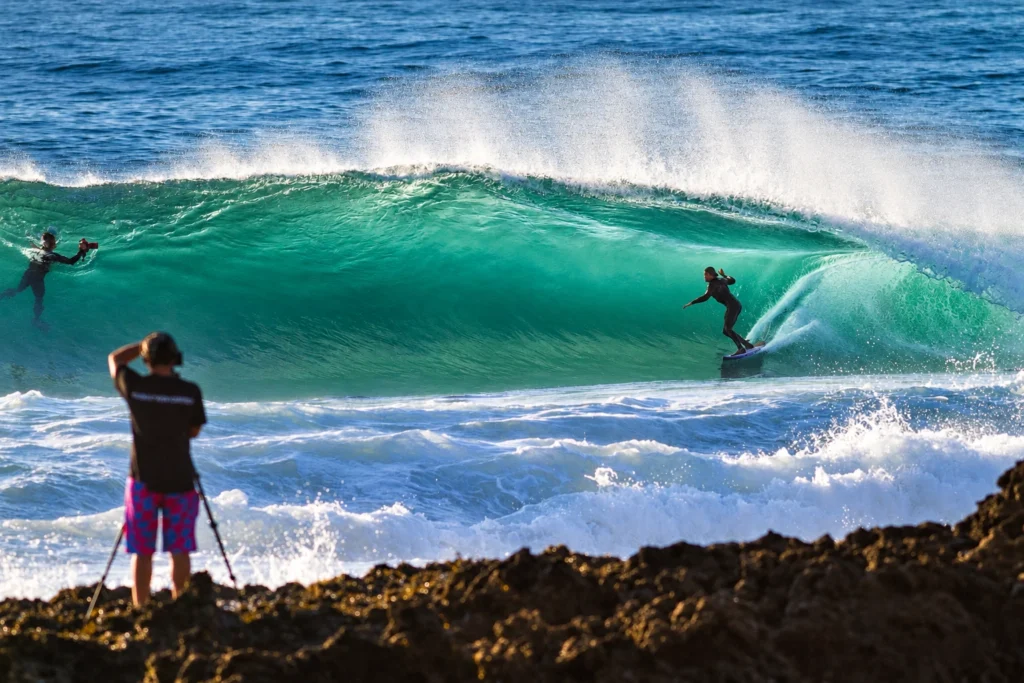 Surfista dentro de um tubo em onda verde, observado por fotógrafo sobre as pedras